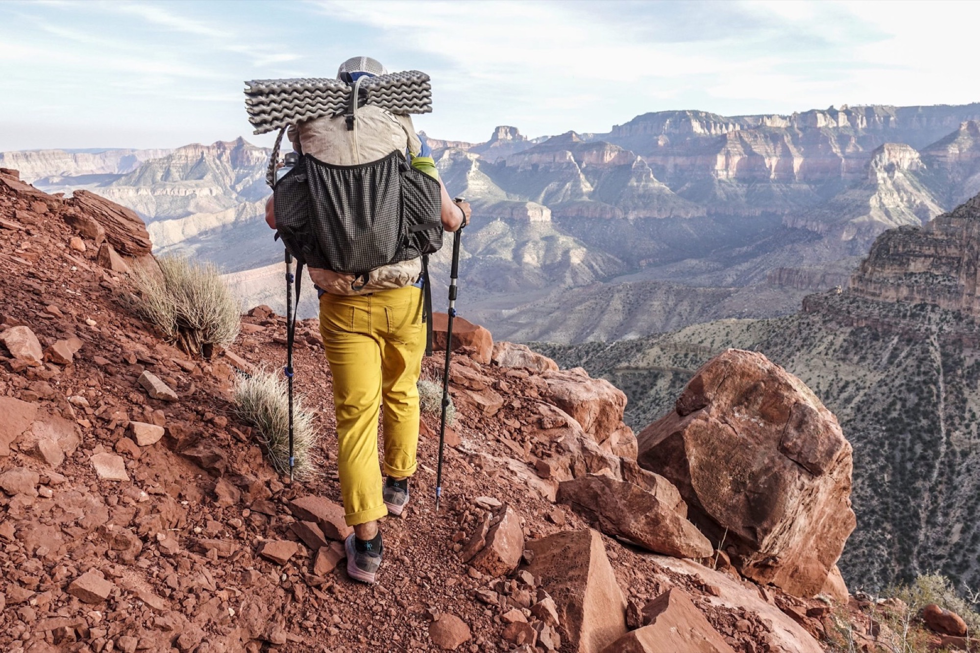 A backpacker in yellow pants hiking down a red rock trail into a canyon