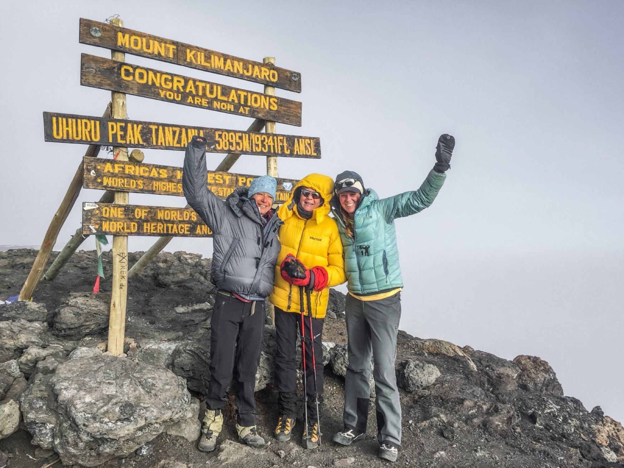Three women cheering at the Uhuru Peak summit sign on Kilimanjaro