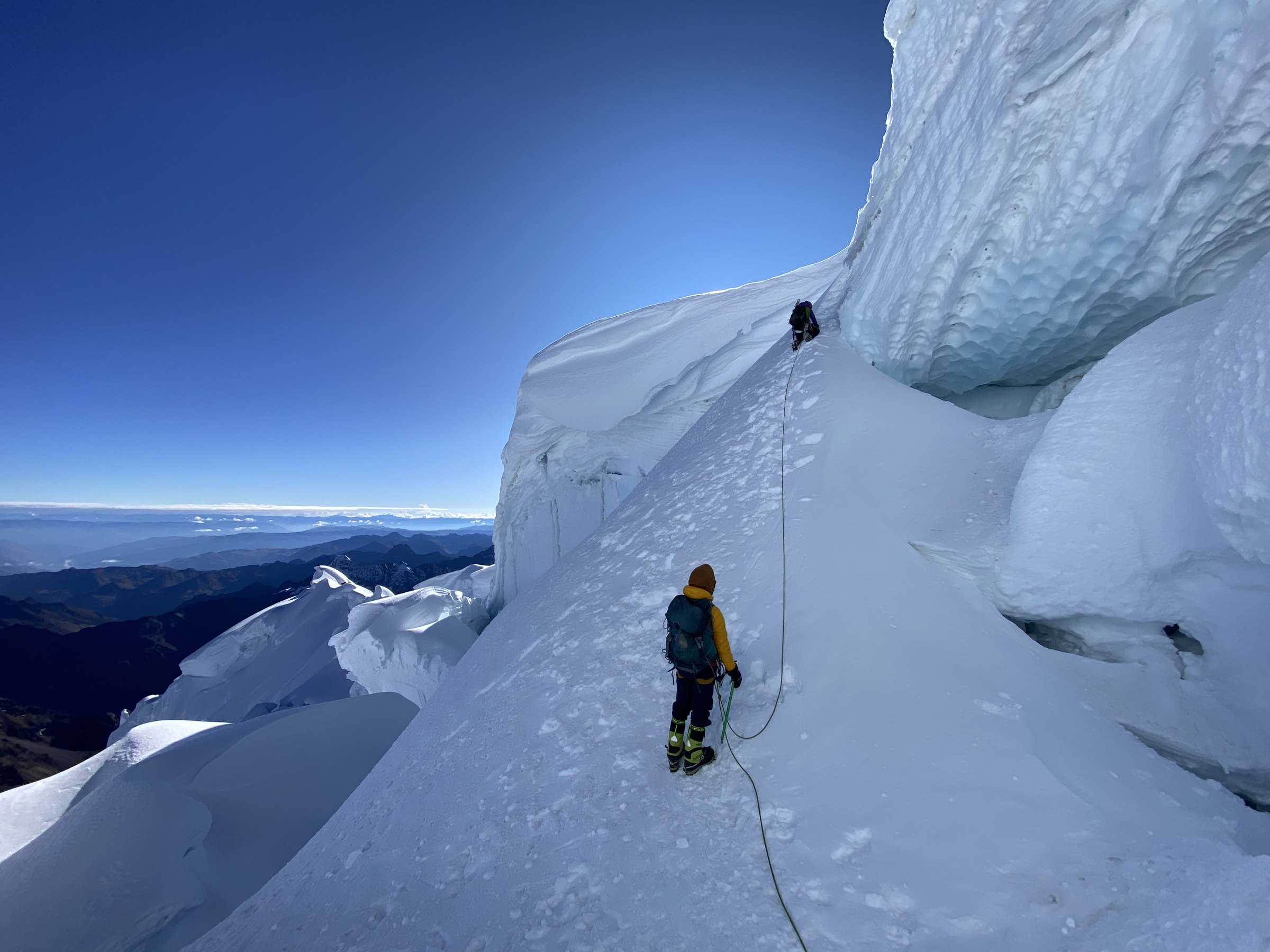A roped climber in a yellow jacket below a steep snow ramp as her partner leads up past an ice cliff in the Cordillera Blanca